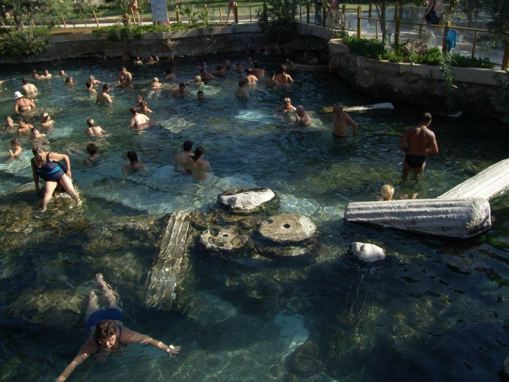 부분적으로 물에 잠긴 고대 기둥과 돌이 있는 자연 수영장에서 수영하는 사람들. 이 지역은 녹지로 둘러싸여 있습니다.