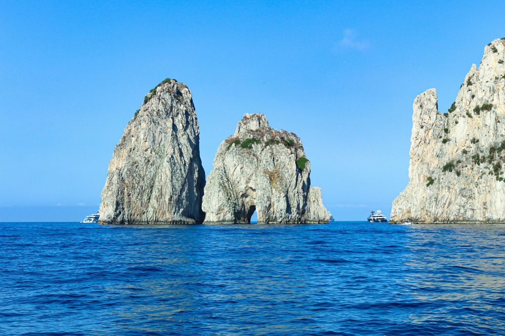 맑고 푸른 하늘 아래 푸른 물로 둘러싸인 커다란 바위 세 개에 초목이 쌓여 있습니다. 인근에 보트가 보입니다.