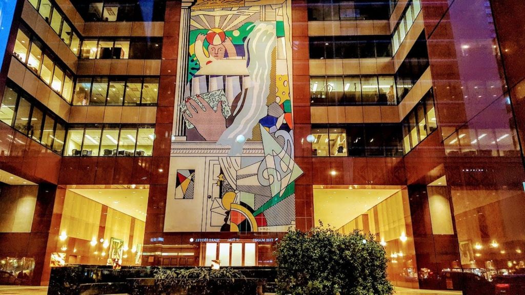 건물 외관에 추상적인 예술품이 있는 커다란 다채로운 벽화로, 조명이 있는 창문과 녹지가 전면에 있습니다.