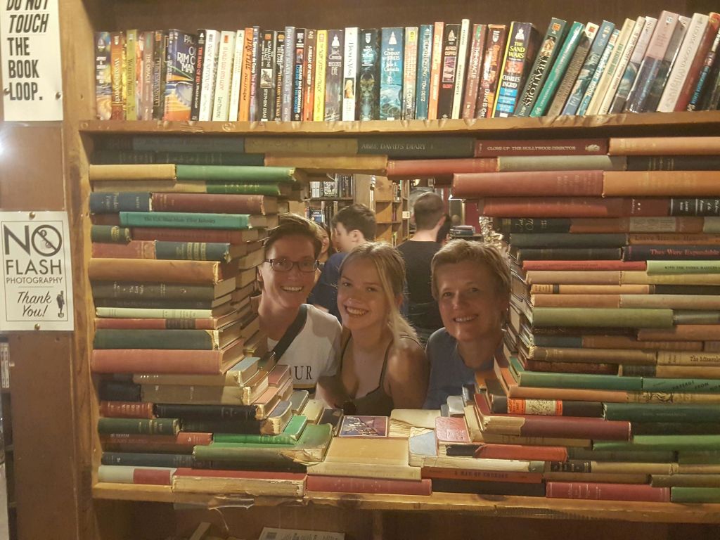 세 사람이 책장을 가득 채우고 더 많은 사람들이 둘러보는 배경과 함께 겹겹이 쌓인 책으로 만든 구멍을 통해 미소를 짓고 있습니다.