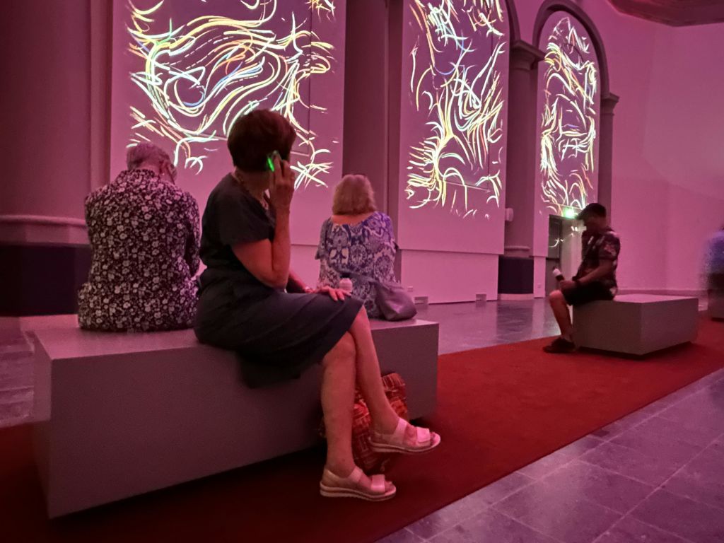 벽에 추상적인 불빛이 투영된 어두운 방의 벤치에 앉아 있는 사람들. 한 사람이 전화를 사용하고 있습니다.