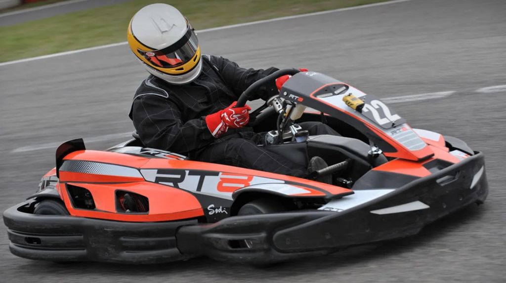 트랙에서 주황색과 검은색 고카트를 운전하는 레이싱 슈트와 헬멧을 착용한 사람.