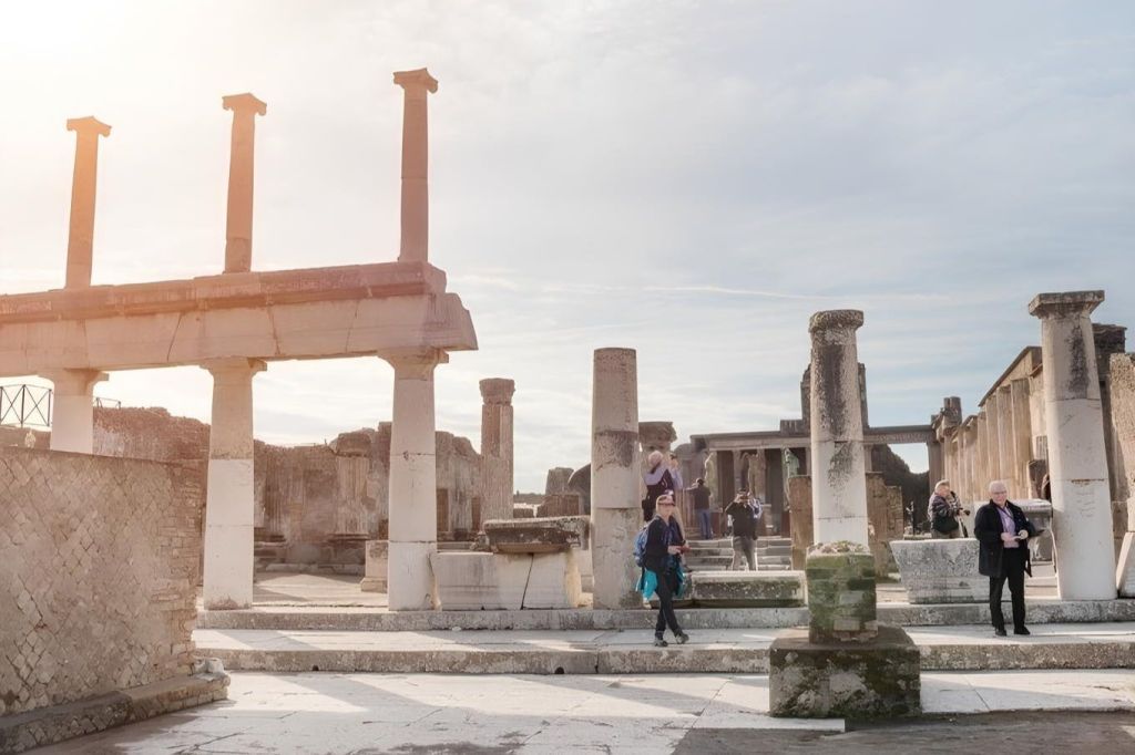 탁한 하늘 아래 석조 기둥과 구조물이 있는 고대 유적 사이를 걷는 사람들.