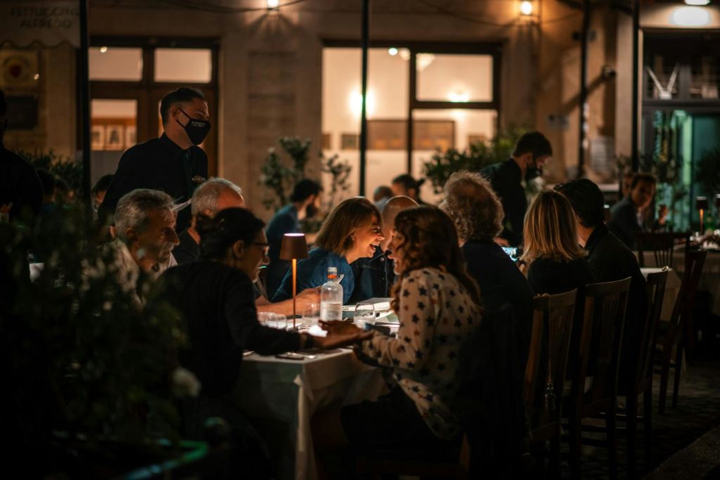 밤에는 야외에서 식사를 즐기고, 마스크를 착용한 게스트를 맞이하고, 부드러운 전등으로 조명이 켜진 테이블을 배경으로 따뜻한 조명이 켜진 건물을 배경으로 식사를 즐겨보세요.