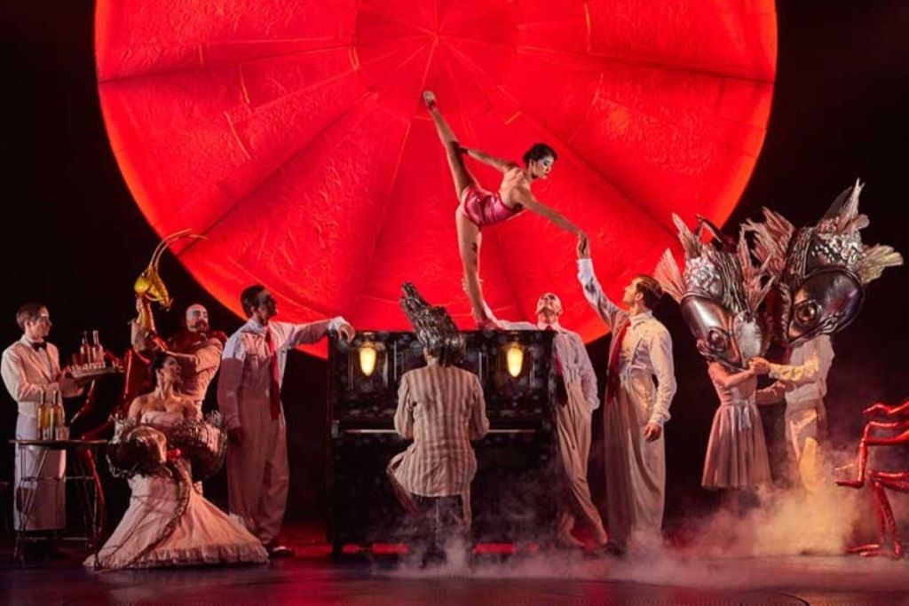 활기찬 빨간색 배경 아래에서 의상을 입은 연주자들과 함께 하는 연극 공연으로, 피아노 위에서 균형을 잡는 댄서가 등장합니다.