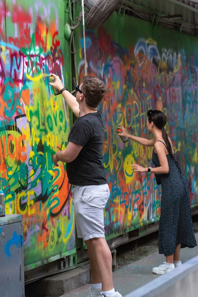 두 사람이 다채로운 색상의 다양한 태그와 디자인으로 덮인 벽에 생동감 넘치는 그래피티를 뿌리고 있습니다.