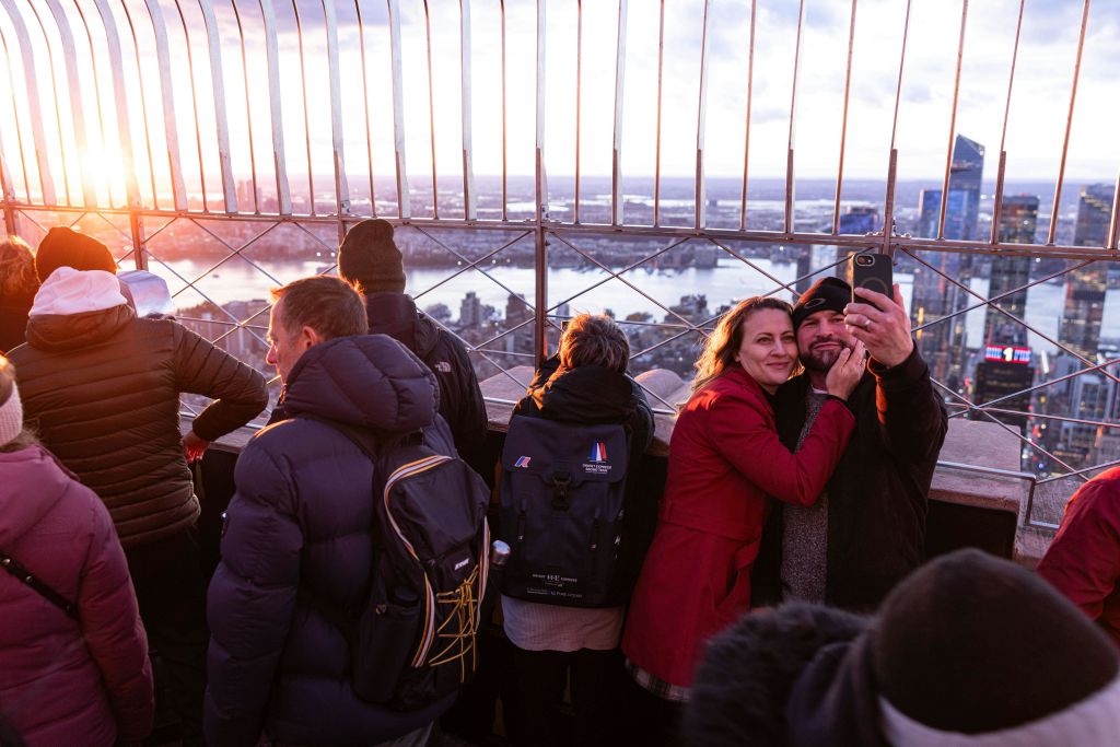 한 커플이 석양을 배경으로 도시 풍경과 강이 보이는 전망대에서 셀카를 찍고 있습니다.