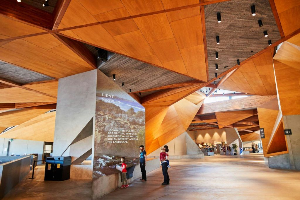 기하학적 목재 천장판이 있는 현대적인 건물 내부에 세 사람이 있습니다. 한 사람이 퍼스트 피플 (First Peoples) 에 대한 벽 디스플레이를 읽습니다.