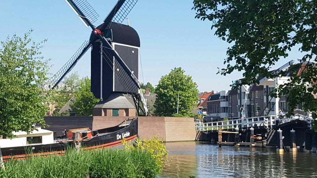 청명한 하늘 아래 녹음과 건물로 둘러싸인 "De Liefde" 라는 이름의 계류 보트가 있는 운하 옆에 풍차가 있습니다.