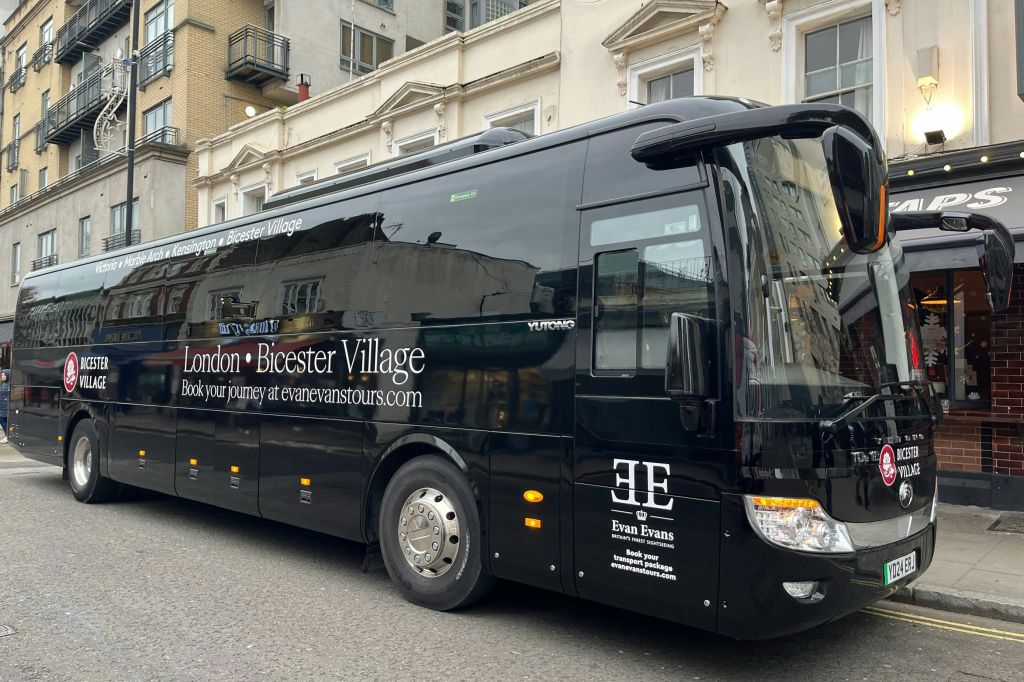 베이지색 건물 앞 거리에 주차된 에반 에반스 투어 (Evan Evans Tours) 를 위한 검은색 투어 버스가 런던에서 비스터 빌리지까지 운행합니다.