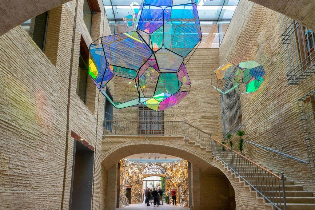 벽돌 벽, 화려한 기하학적 매달린 조각, 화려한 아치형 통로를 걷는 사람들이 있는 현대적인 아트리움입니다.