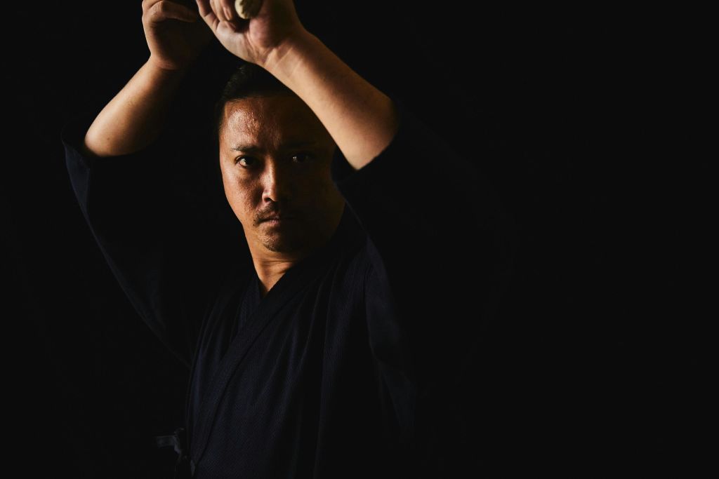 검은색 옷을 입은 사람이 검은색 배경으로 머리 위로 검을 들고 집중된 표정을 짓고 있습니다.