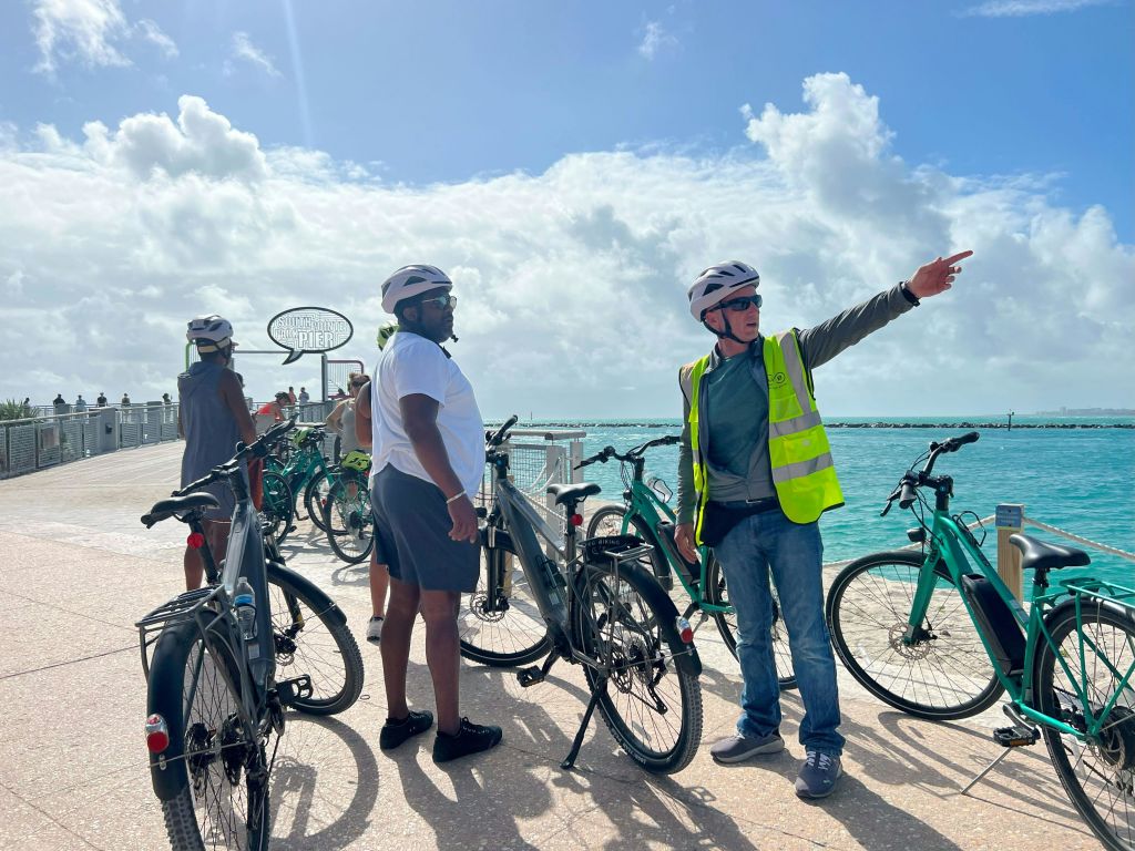 헬멧과 안전 조끼를 착용한 두 남성이 바닷가 대여 자전거 근처에 서 있습니다. 한 남자가 지평선을 가리켰다.