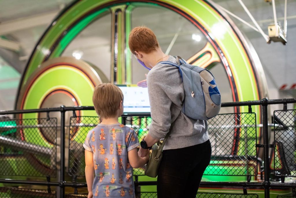 커다랗고 화려한 색상의 기계가 배경으로 놓인 디스플레이를 바라보며 한 여성과 어린이가 손을 맞잡고 서 있습니다.