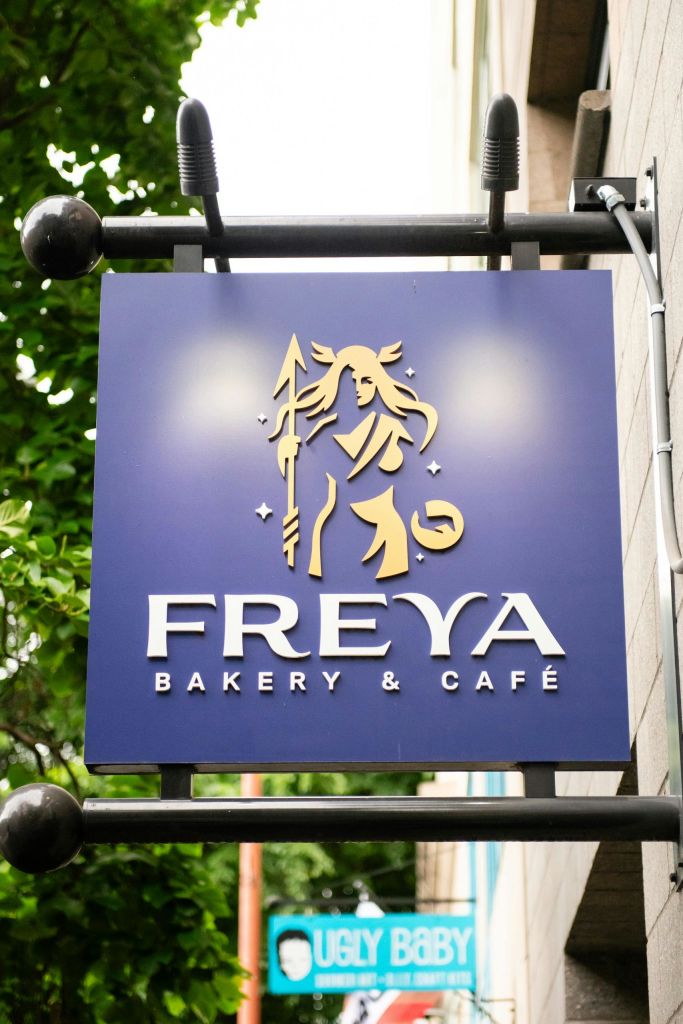 푸른색 배경에 금색 요소가 특징인 창을 들고 있는 스타일리시한 인물이 있는 Freya Bakery & Café의 간판입니다.