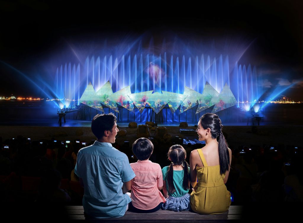 4명의 가족이 밤에는 분수와 조명 구조물을 배경으로 활기찬 물과 빛의 쇼를 관람합니다.