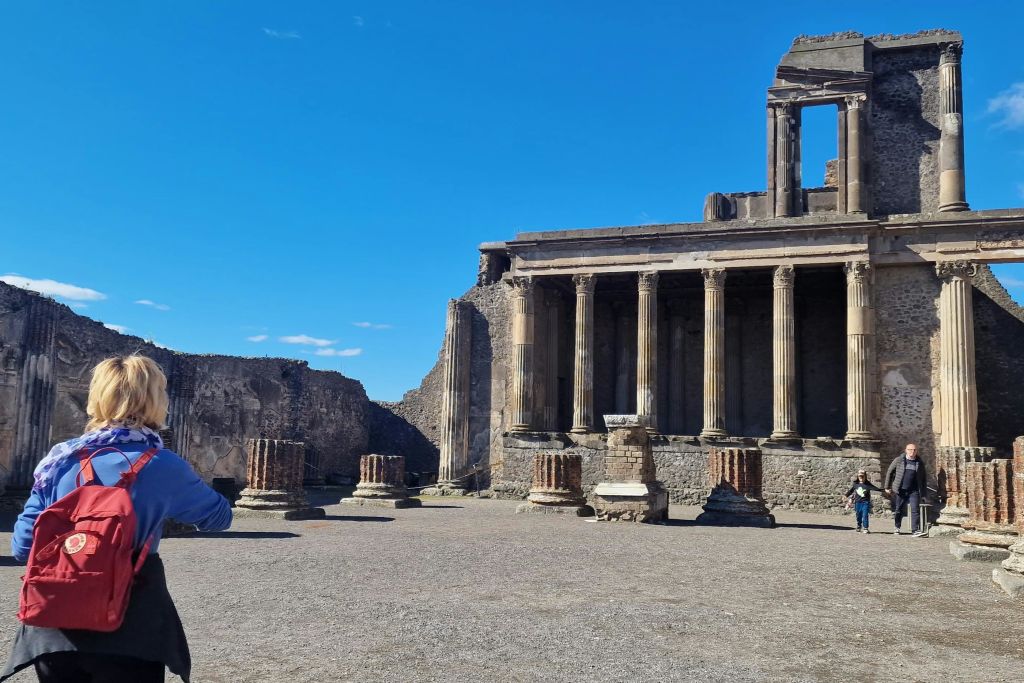 맑고 푸른 하늘 아래 기둥과 부분적으로 서 있는 건물이 있는 고대 유적지로, 장면 왼쪽에 사람이 있습니다.