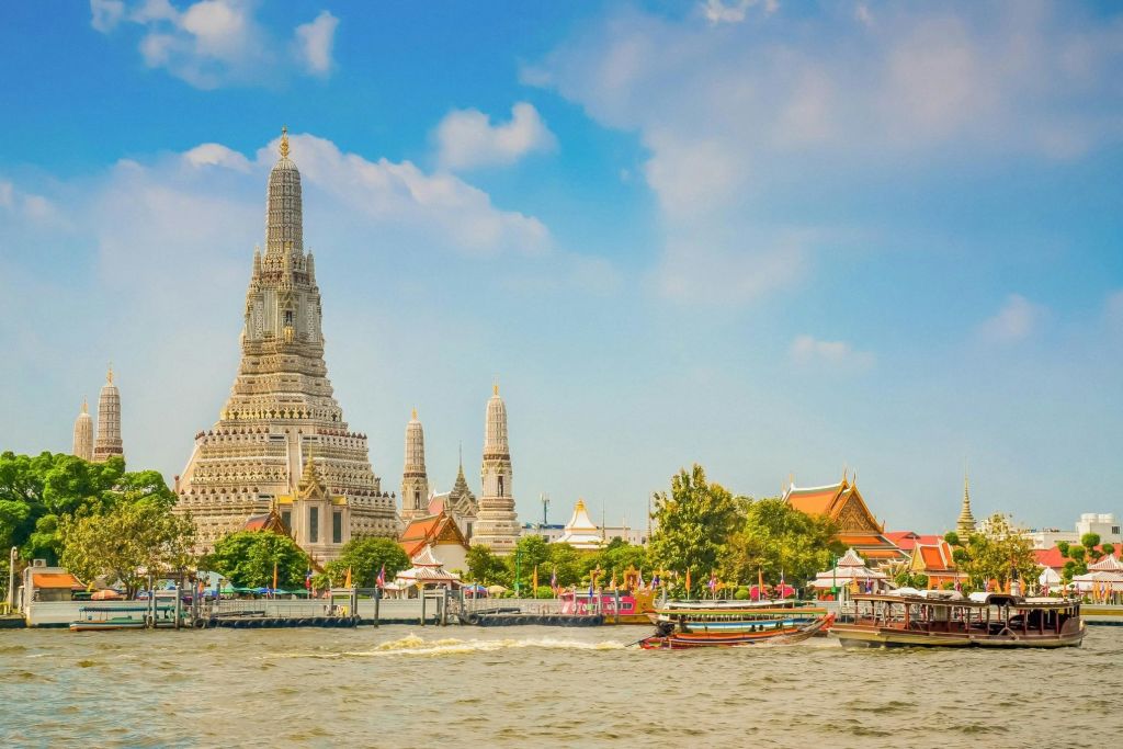 높은 첨탑, 형형색색의 옥상, 푸른 하늘 아래 물 위의 보트가 있는 왓 아룬 사원의 강변 풍경.