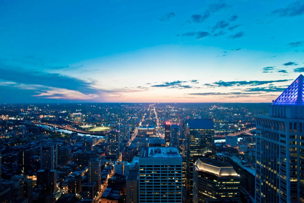 조명이 켜진 건물, 거리, 부분적으로 흐린 하늘 아래 강이 있는 석양의 도시 경관을 조망할 수 있습니다.