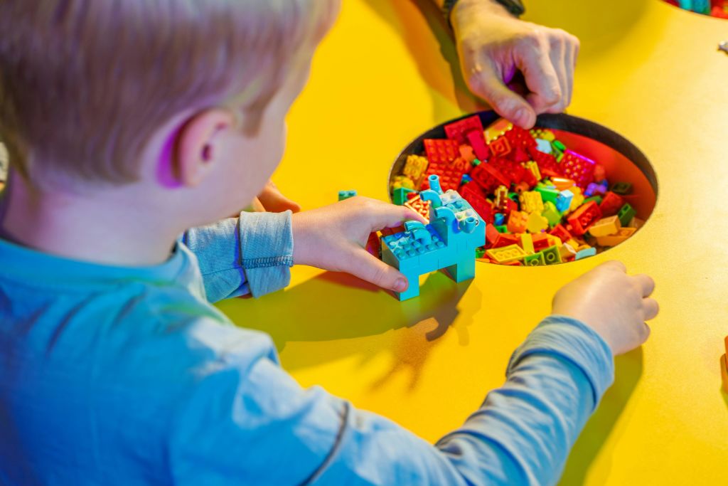 어린이는 노란색 테이블에 파란색과 녹색 레고 벽돌을 쌓고 어른의 손은 중앙의 화려한 벽돌 더미에서 조각을 회수합니다.