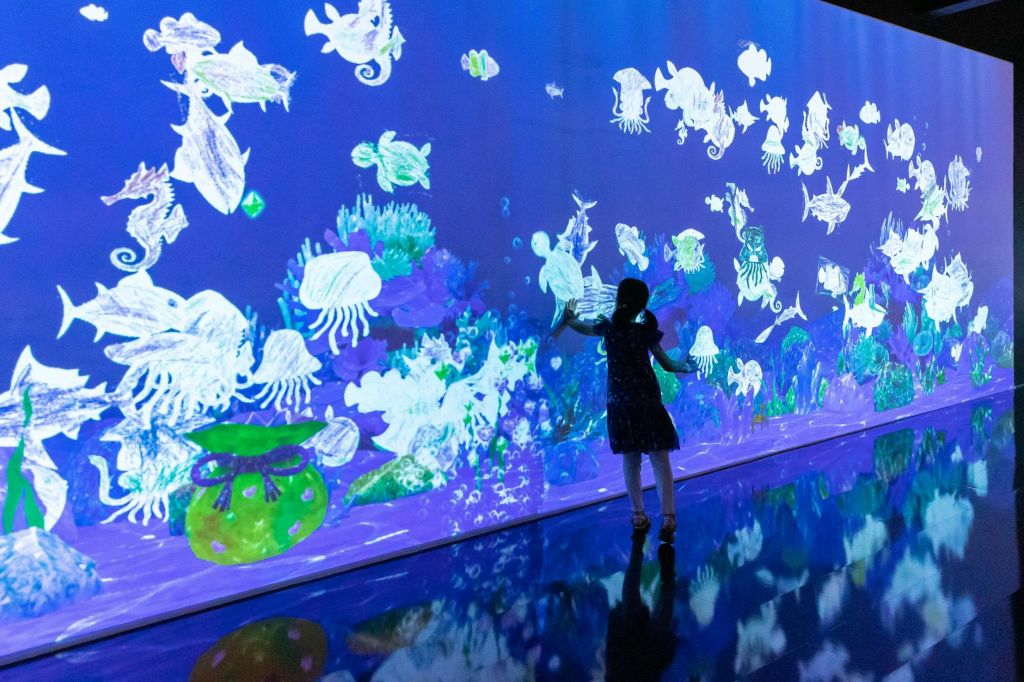 다크 드레스를 입은 사람이 다채로운 해양 생물과 식물을 특징으로 하는 대형 디지털 수중 디스플레이와 상호 작용합니다.
