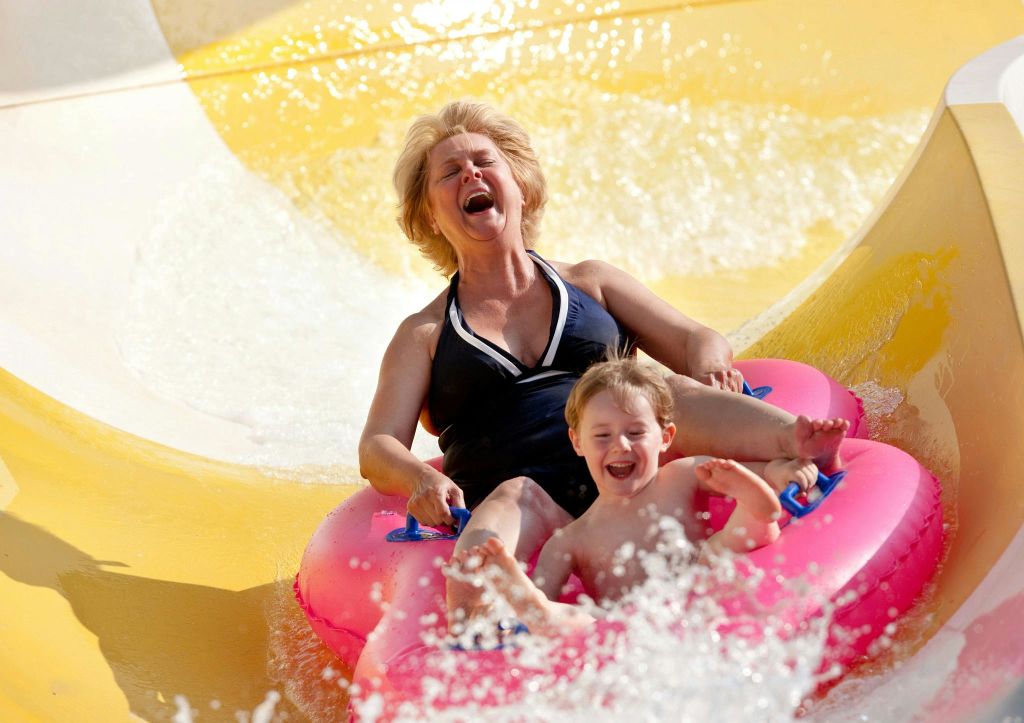 분홍색 팽창식 링을 착용한 여성과 어린이가 워터 슬라이드를 따라 내려가며 웃으며 라이딩을 즐깁니다.