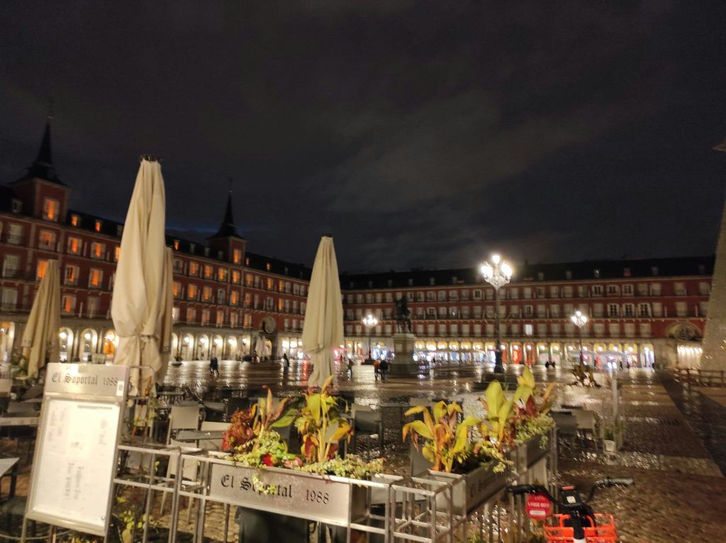 조명이 켜진 전등 기둥, 닫힌 우산, 화분, 중앙에 동상이 있는 광장의 야경, 건물을 배경으로 한 건물.