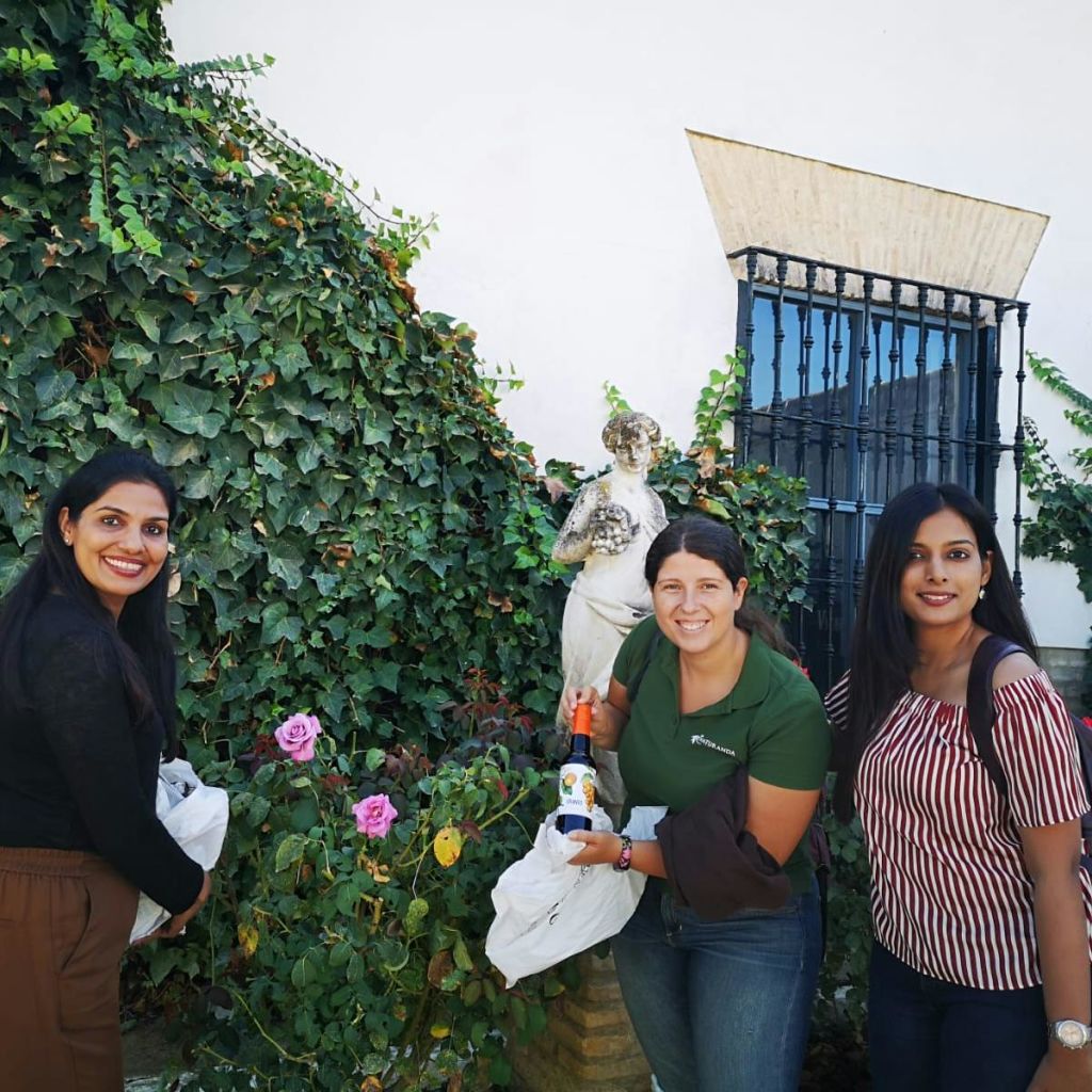 아이비나무와 꽃이 피어 있는 정원 동상 근처의 흰 벽에 걸린 창문 앞에서 세 명의 여성이 포즈를 취하고 있습니다.