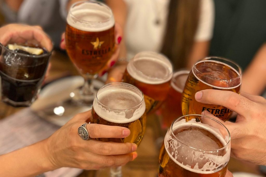 사람들은 손과 팔이 보이는 토스트에 에스트렐라 맥주를 넣고 나무 테이블을 둘러싸고 칵테일을 마십니다.