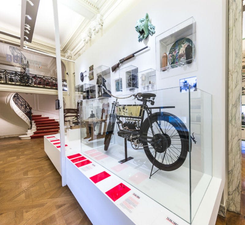 유리 케이스에 빈티지 오토바이, 벽에 걸린 역사적 유물, 계단을 배경으로 한 박물관 전시물.