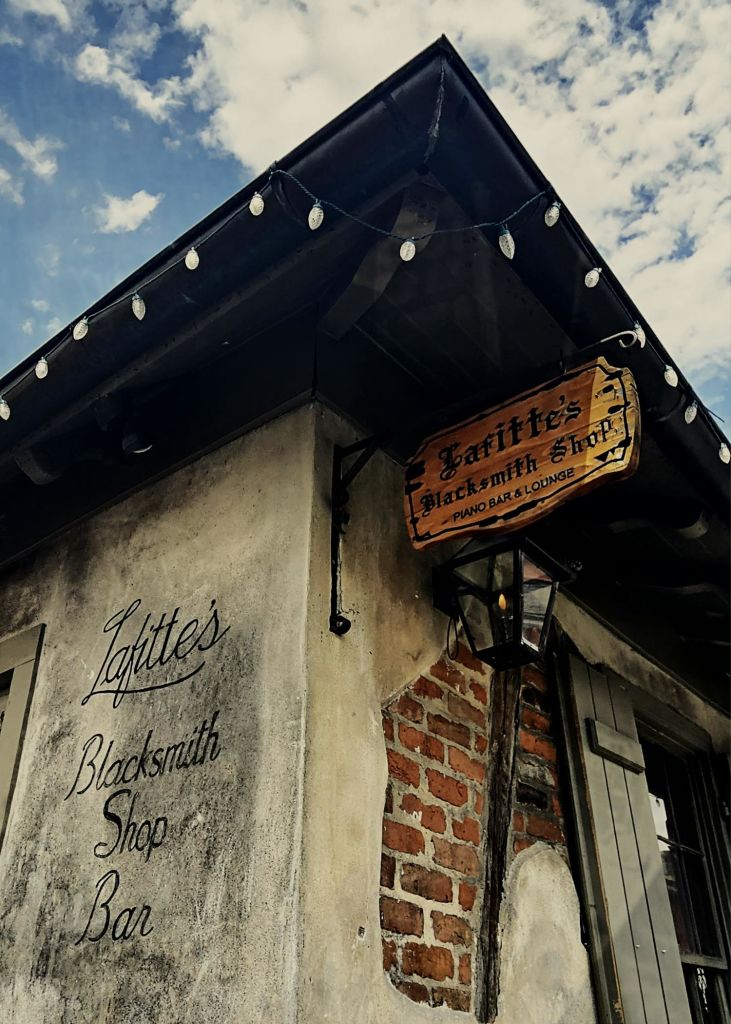 "Lafitte's Blacksmith Shop" 이라는 간판이 달린 유서 깊은 건물의 외관, 불빛의 끈, 푸른 하늘.