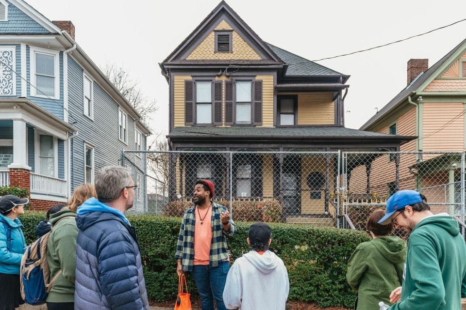 파란색 주택이 인접한 울타리가 있는 2층짜리 노란색 주택 앞에서 한 남성이 소그룹에 대해 이야기하고 있습니다.
