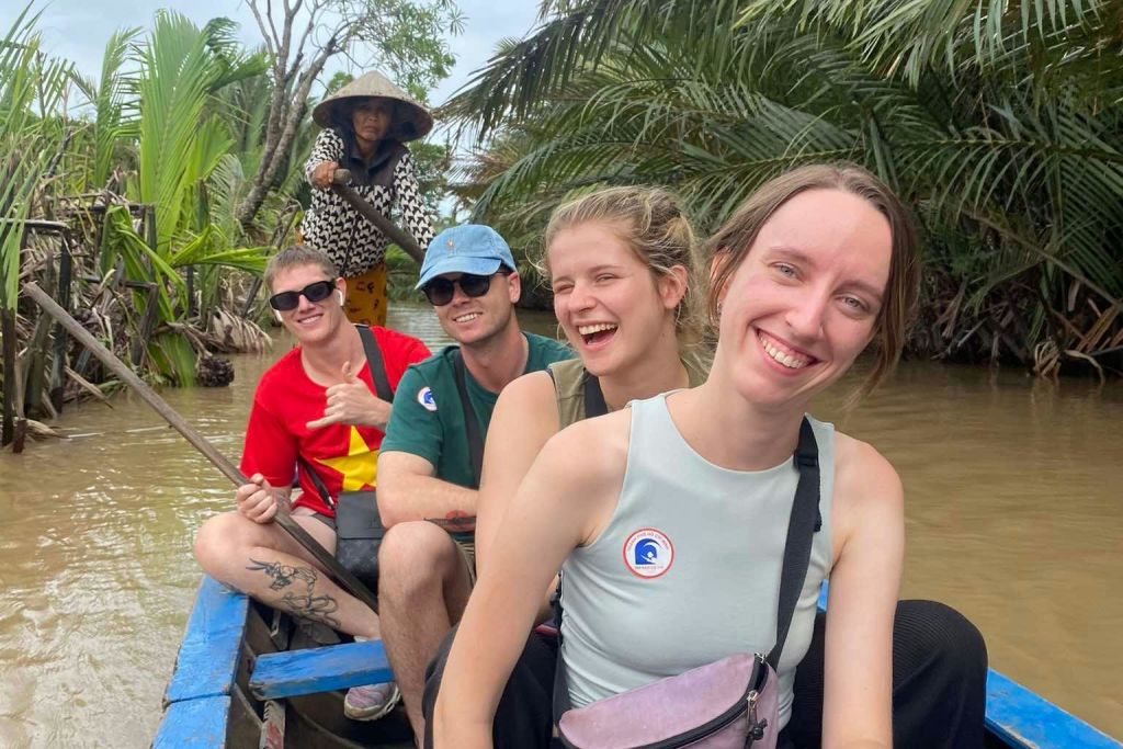 강가에서 보트를 타고 미소를 짓고 있는 네 사람. 원뿔 모양의 모자를 쓴 한 사람이 울창한 녹음 속에서 배경에 서서 노를 젓고 있습니다.