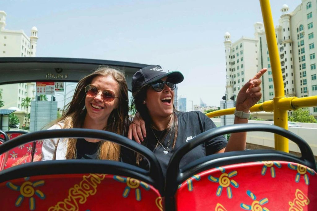 빨간색 오픈탑 투어 버스에 앉아 도시 건물과 맑은 하늘을 배경으로 웃고 있습니다.