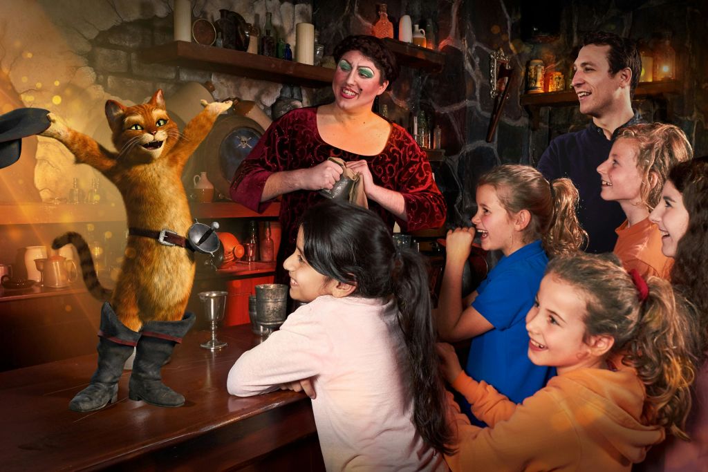 중세 의상을 입은 한 여성이 소박한 분위기의 화려한 옷을 입고 미소 짓는 남성과 어린이 그룹과 상호 작용합니다.