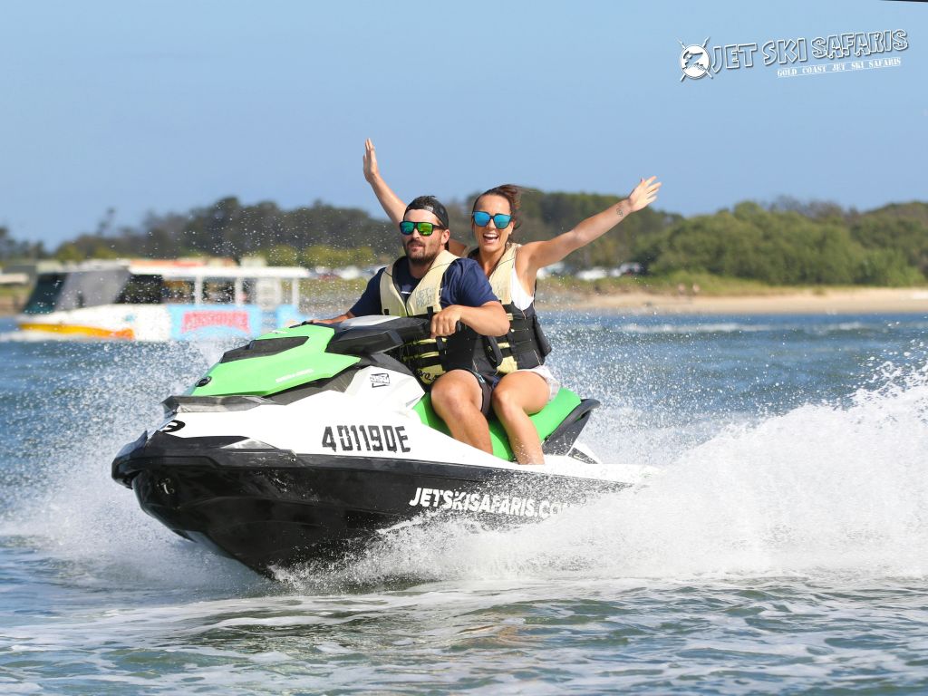 구명 조끼를 입은 두 사람이 물 위에서 제트 스키를 타고 있습니다. 뒤쪽의 여자가 팔을 들어 올렸다. 배경에는 보트와 나무가 있습니다.