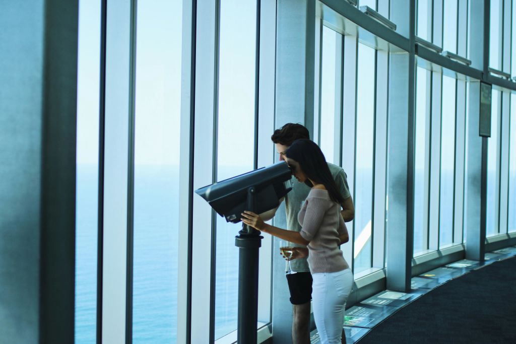 바다가 내려다보이는 커다란 창문이 있는 실내 전망대에서 쌍안경으로 망원경을 들여다보는 커플.