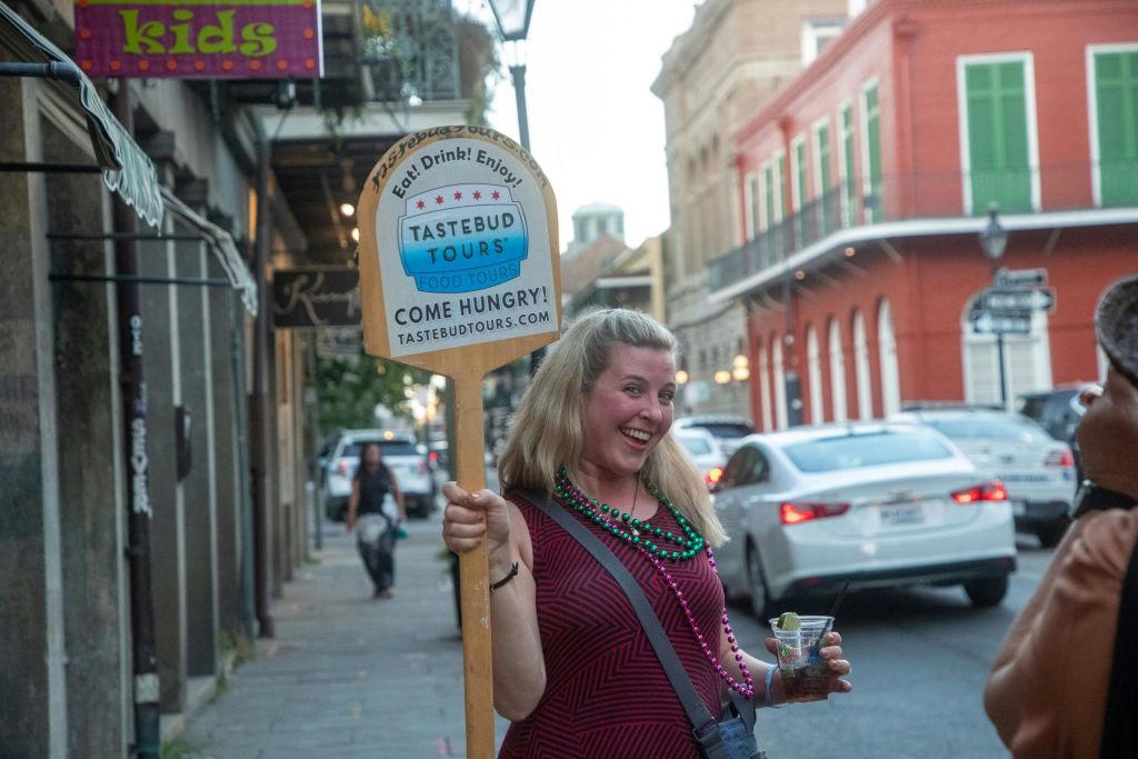 화려한 건물과 자동차를 배경으로 활기찬 거리에서 "타스테부드 투어" 표지판을 들고 웃고 있는 여성.