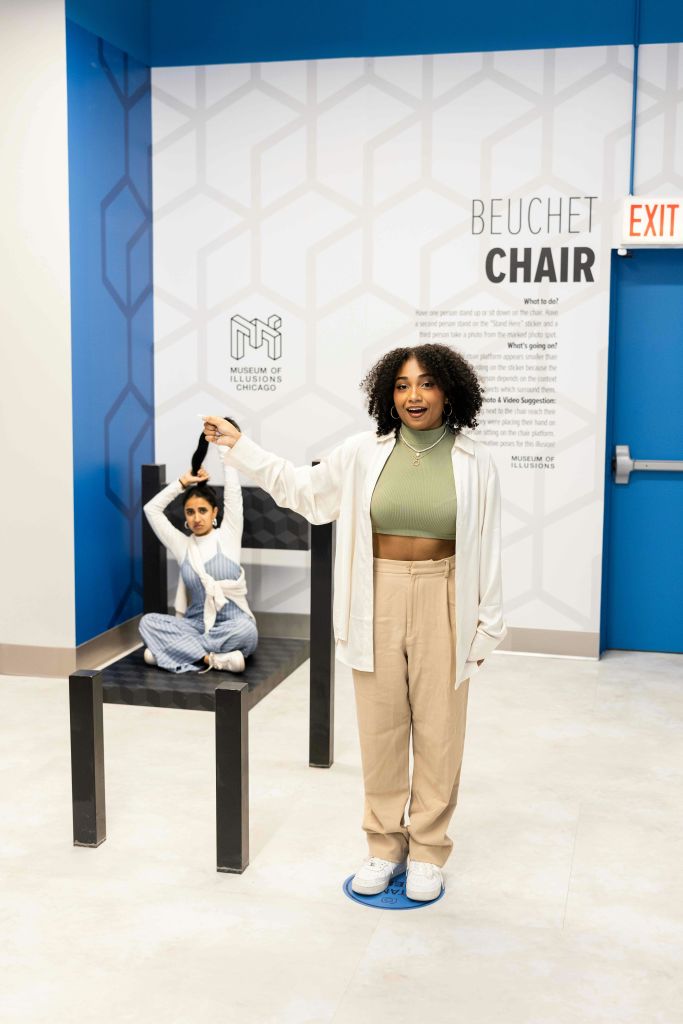 환상의 두 여성이 전시되어 있습니다. 한 여성은 의자 소품에 작게 보이고, 다른 한 여성은 의자 위에 손을 들고 정면에 서 있습니다.