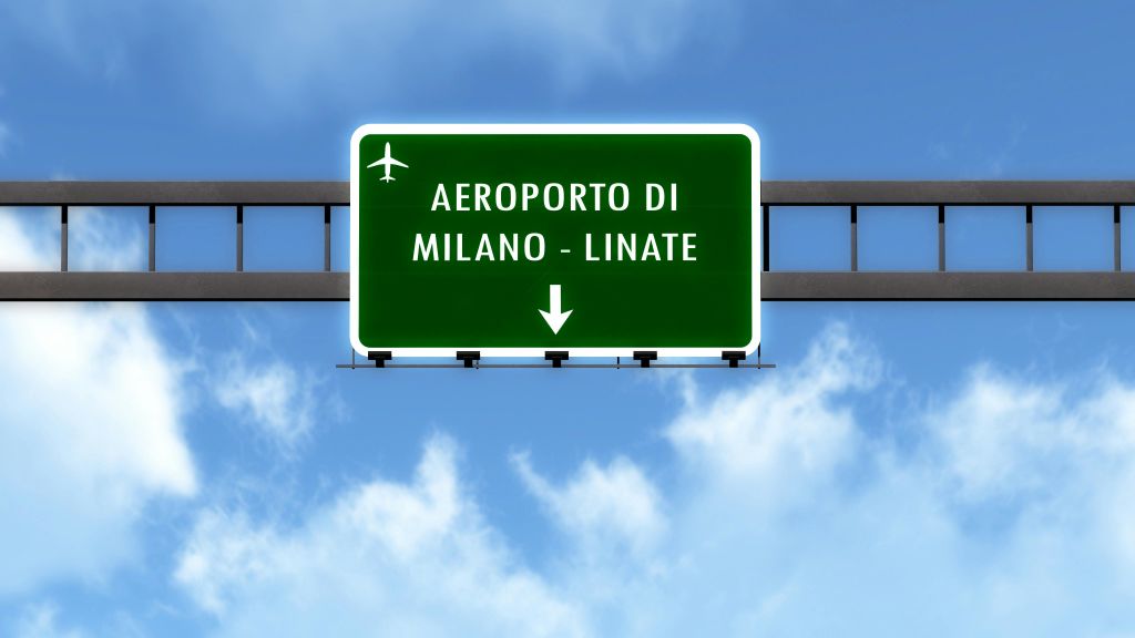 "AEROPORTO DI MILANO - LINATE" 텍스트가 있는 녹색 도로 표지판과 구름이 있는 푸른 하늘을 배경으로 한 비행기 기호.