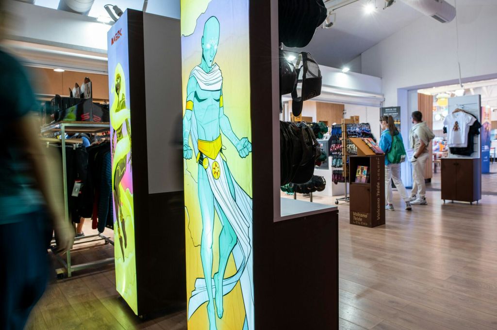 파란색 근육질 캐릭터의 커다란 포스터와 하얀 망토가 진열된 상점. 일부 쇼핑객은 백그라운드에서 볼 수 있습니다.