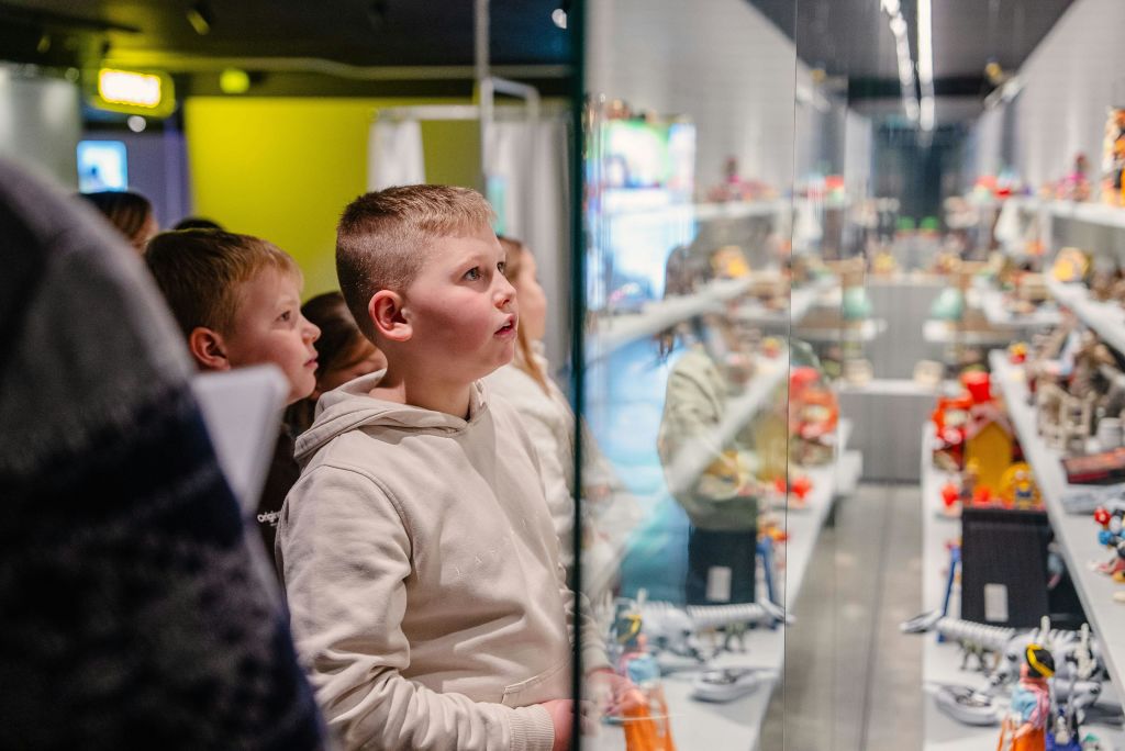 후드티를 입은 어린이들은 사람들에게 둘러싸인 조명이 밝은 실내 전시물에 전시된 것을 세심하게 관찰합니다.