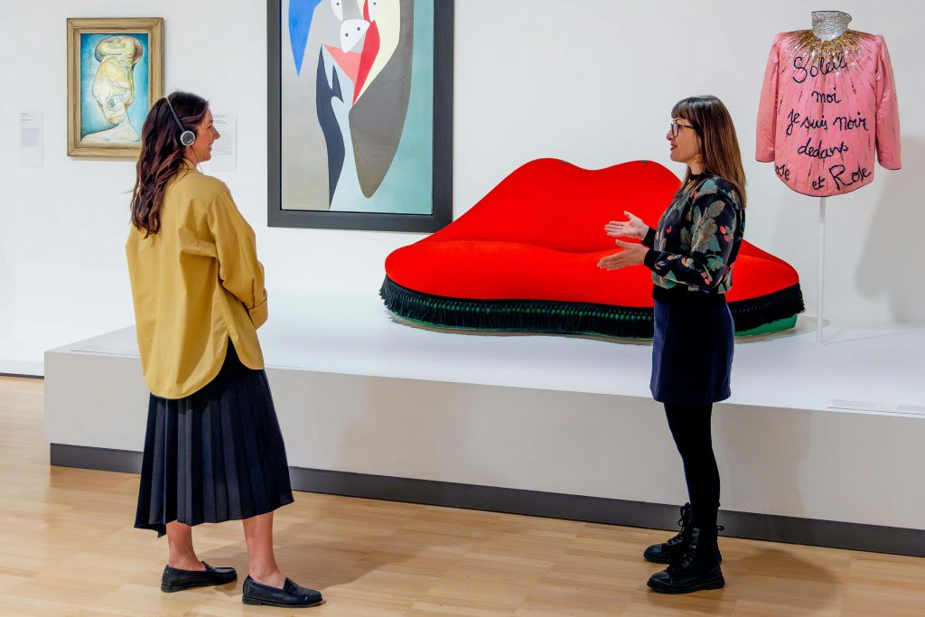 미술 전시장에서 한 여성이 빨간색 입술 모양의 소파와 분홍색 장식 재킷 앞에 서서 제스처를 취하고 있습니다.