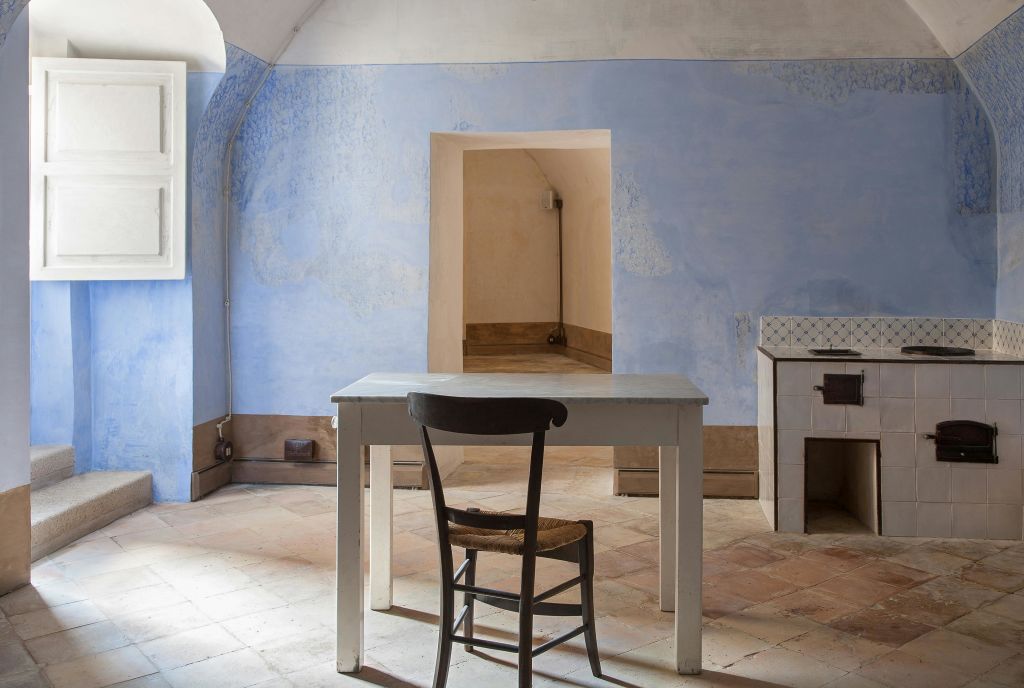 밝은 파란색 벽, 흰색 테이블, 타일 바닥에 나무 의자가 있는 심플한 객실입니다. 열린 출입구와 창문이 보입니다.