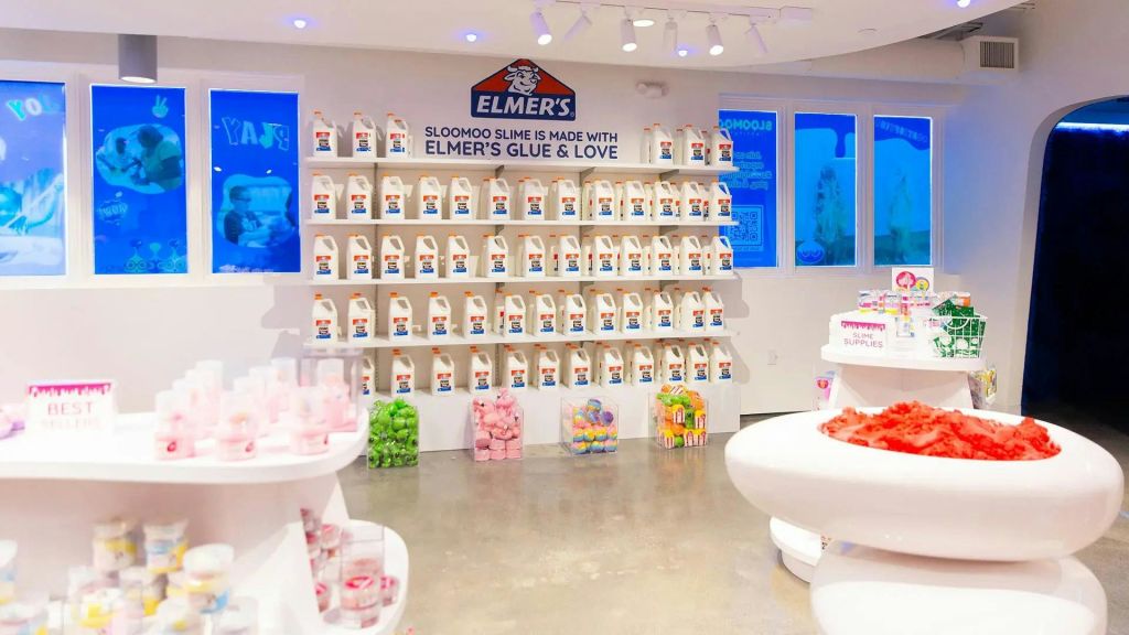 흰색 선반에 다양한 다채로운 점액질 제품이 있는 밝은 조명 매장의 선반에 배치된 엘머의 접착제 병이 전시되어 있습니다.