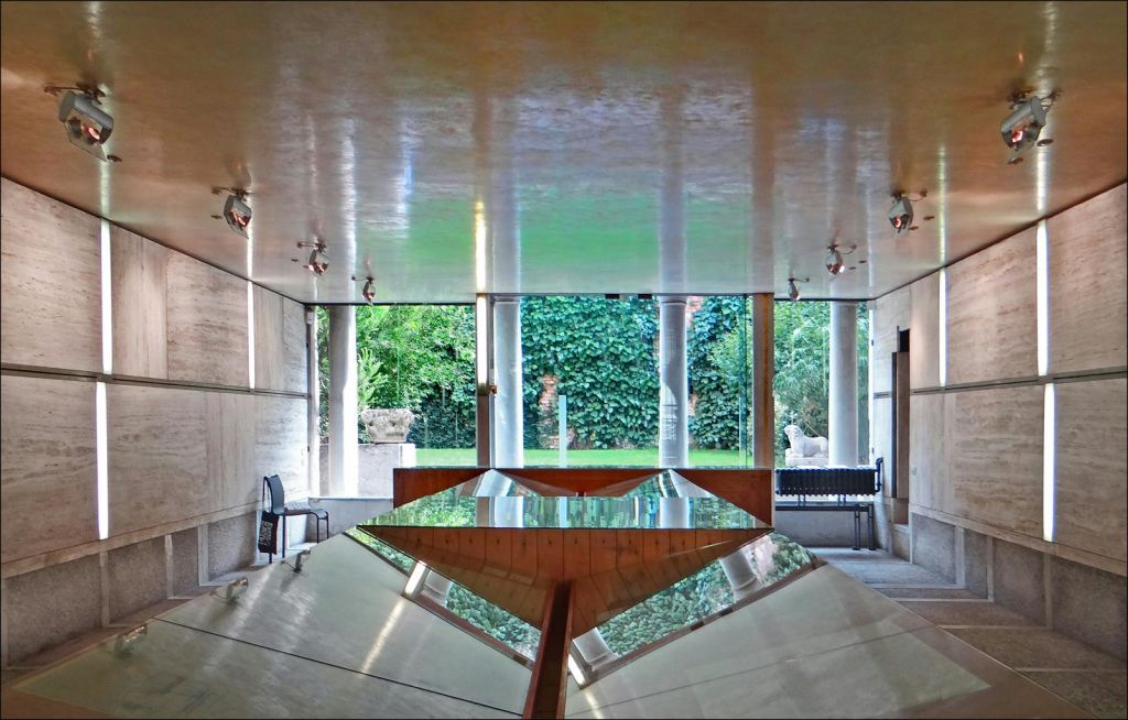반사되는 표면과 나무와 조각상이 있는 울창한 정원으로 통하는 커다란 창문이 있는 현대적인 객실입니다.