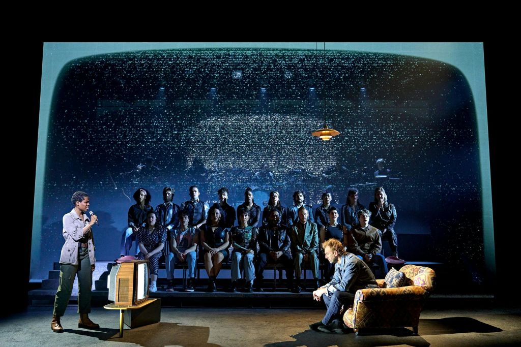 오래된 텔레비전 옆에 앉아 마이크를 잡으며 무대 뒤에 앉아 있는 한 남성.