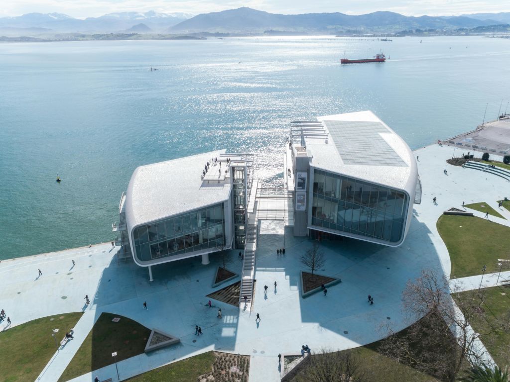 바닷가 옆의 커다란 유리창이 있는 현대적이고 분할된 건물, 포장된 지역을 걷는 사람들, 산을 배경으로 한 건물입니다.