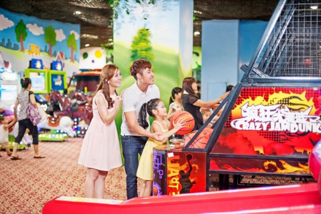 남자, 여자, 어린이를 포함한 가족이 밝은 실내 놀이 시설에서 아케이드 농구 게임을 하고 있습니다.