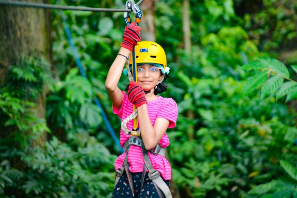 노란색 헬멧과 분홍색 셔츠를 입은 어린이가 울창하고 푸른 숲에서 집라인 하네스를 잡고 있습니다.